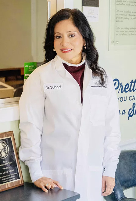 The image shows a person standing behind a desk, wearing a white lab coat with a name tag, smiling at the camera.