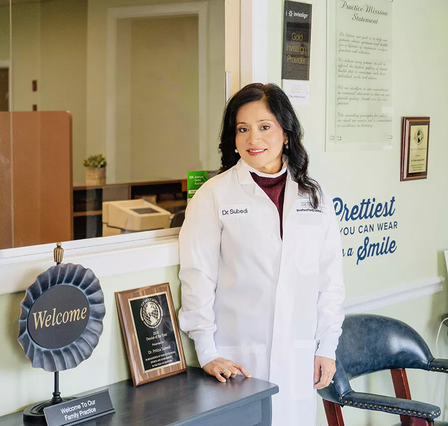 A woman wearing a white lab coat stands behind a desk with a sign that reads  Welcome  and a plaque.