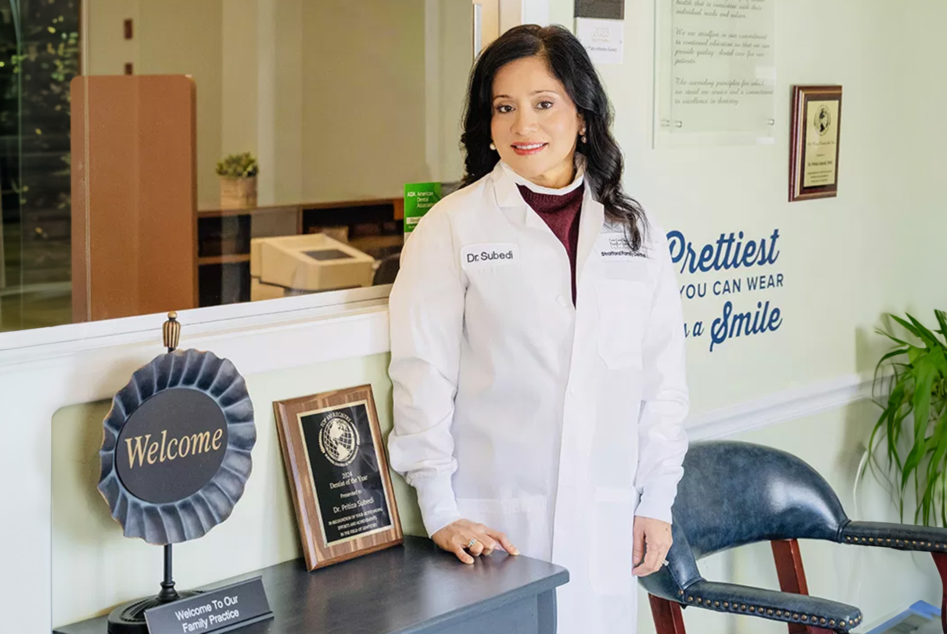 The image depicts a woman wearing a white lab coat standing in an office environment, posing for the camera with a smile.