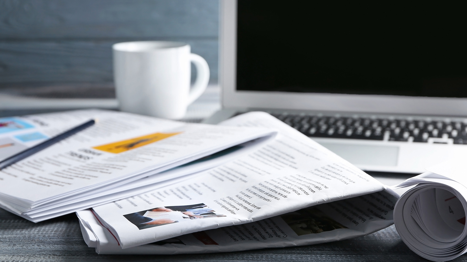 The image shows a collection of newspapers spread out on a desk next to a laptop computer with an open paperback book, suggesting a workspace focused on information gathering or news reporting.