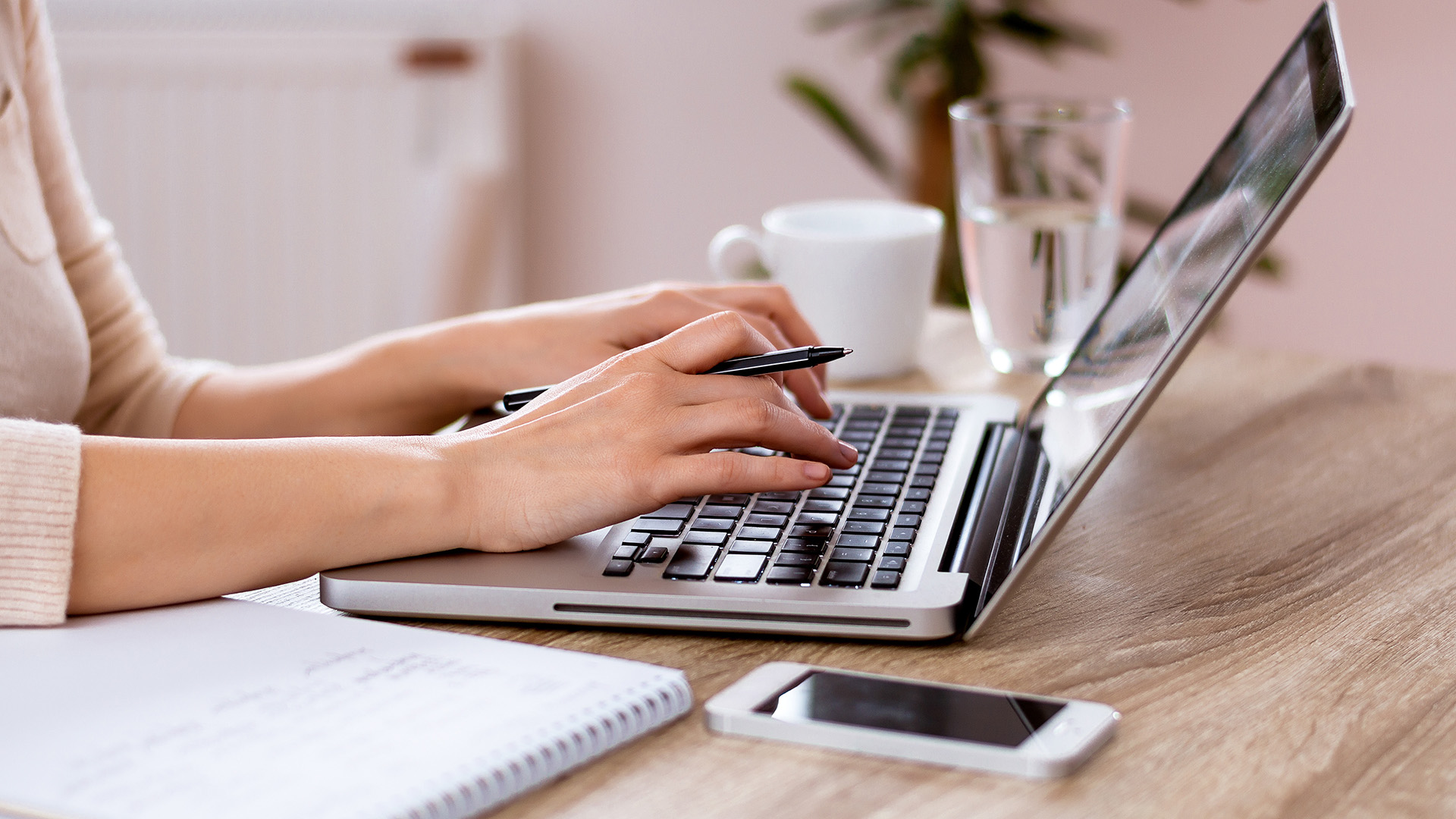 A woman working on her laptop at a desk with a notebook and pen nearby.