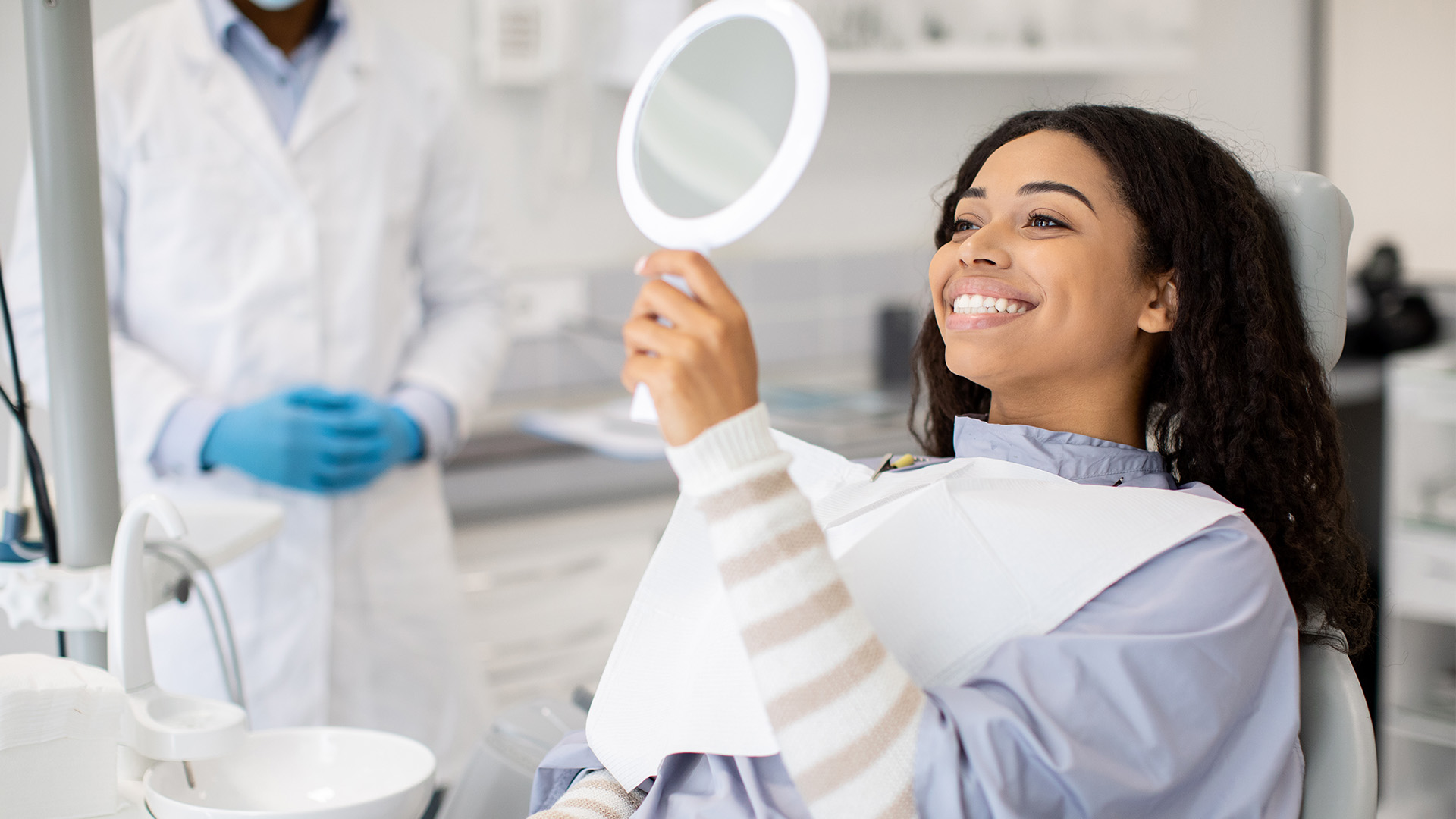 A woman sits in a dental chair, smiling at a mirror with a medical professional standing behind her.