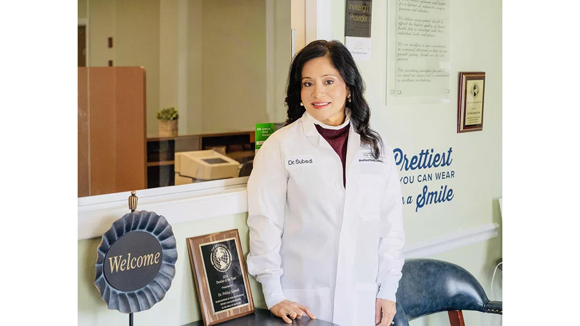 The image shows a person standing in an office environment, wearing a white lab coat, posing for the camera with a smile.