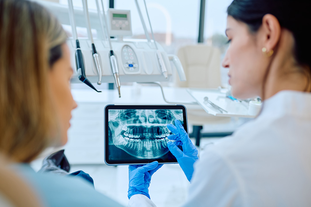 A woman in a lab coat holding up an iPad displaying a 3D model of a human mouth while another person looks on, both in a medical setting with dental equipment around them.