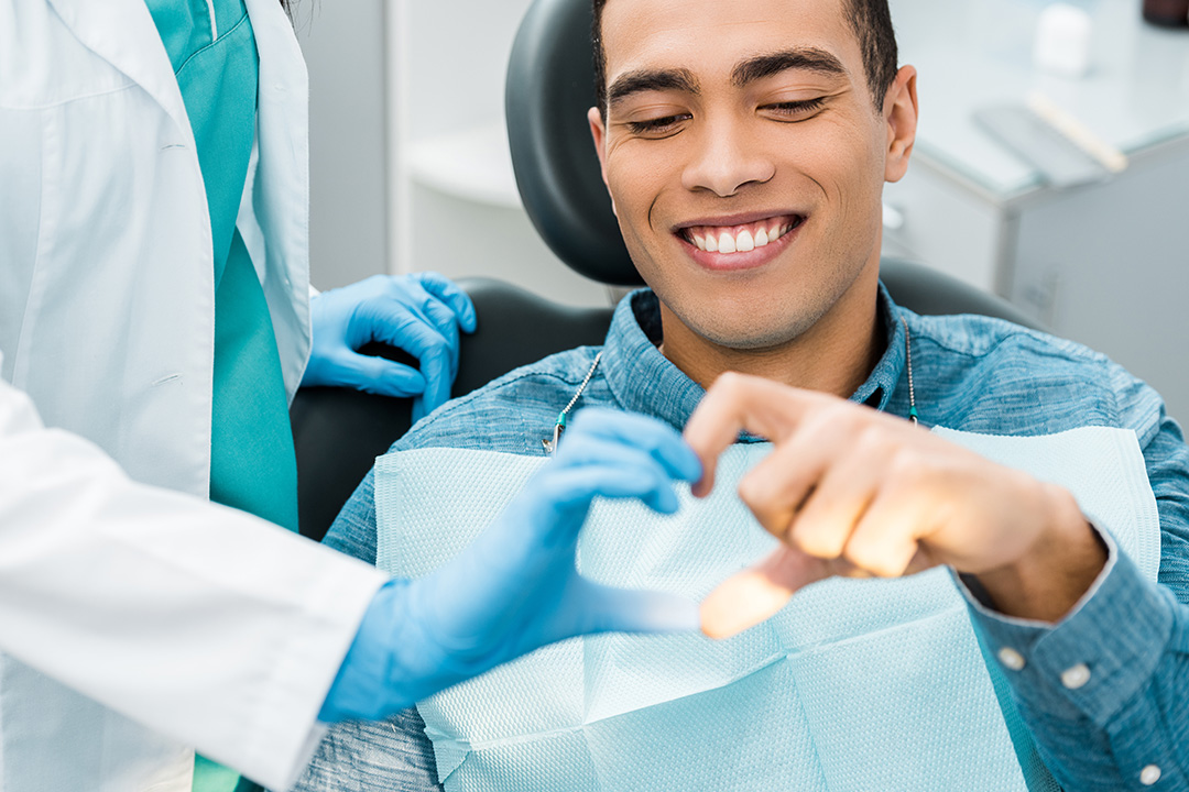 A smiling man wearing blue gloves and sitting in a dental chair while receiving dental care from a professional.