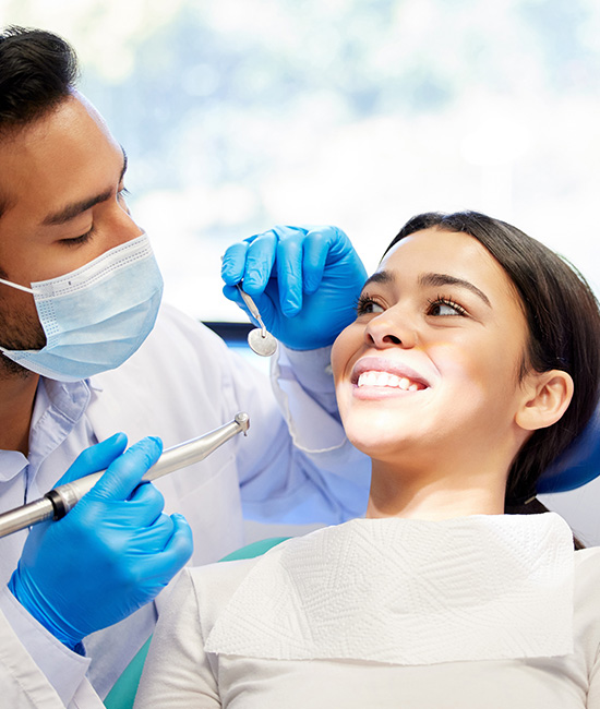 The image shows a dental hygienist performing a cleaning procedure on a patient s teeth, with both individuals wearing masks and gloves for safety during the COVID-19 pandemic.