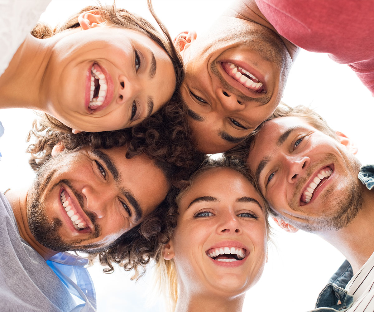 The image shows a group of five individuals smiling at the camera, with their faces arranged around a central point, against a blurred background.