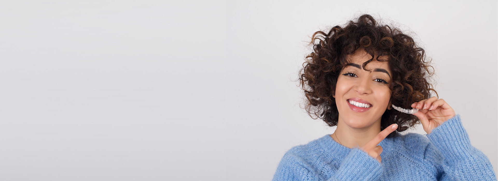 The image shows a person with curly hair smiling while holding a toothbrush, wearing a blue sweater, against a white background.