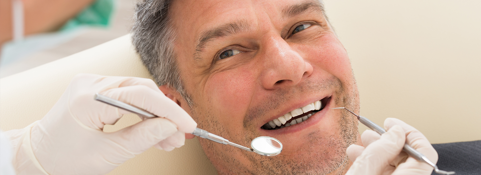 A man sitting in a dental chair with his mouth open while receiving dental care, wearing a white lab coat, with various dental tools visible around him.