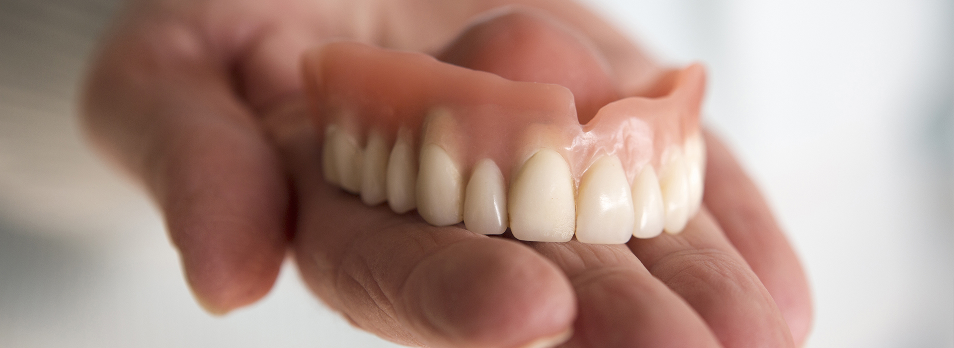 The image shows a person holding an artificial lower denture with multiple teeth, which is placed in their hand against a blurred background.