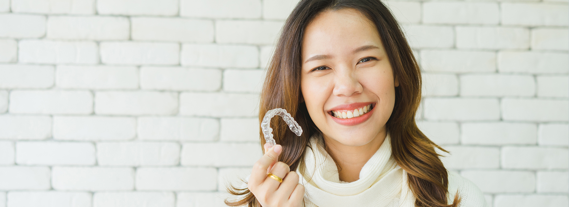A woman smiling at the camera while holding up a toothbrush, set against a brick wall background.
