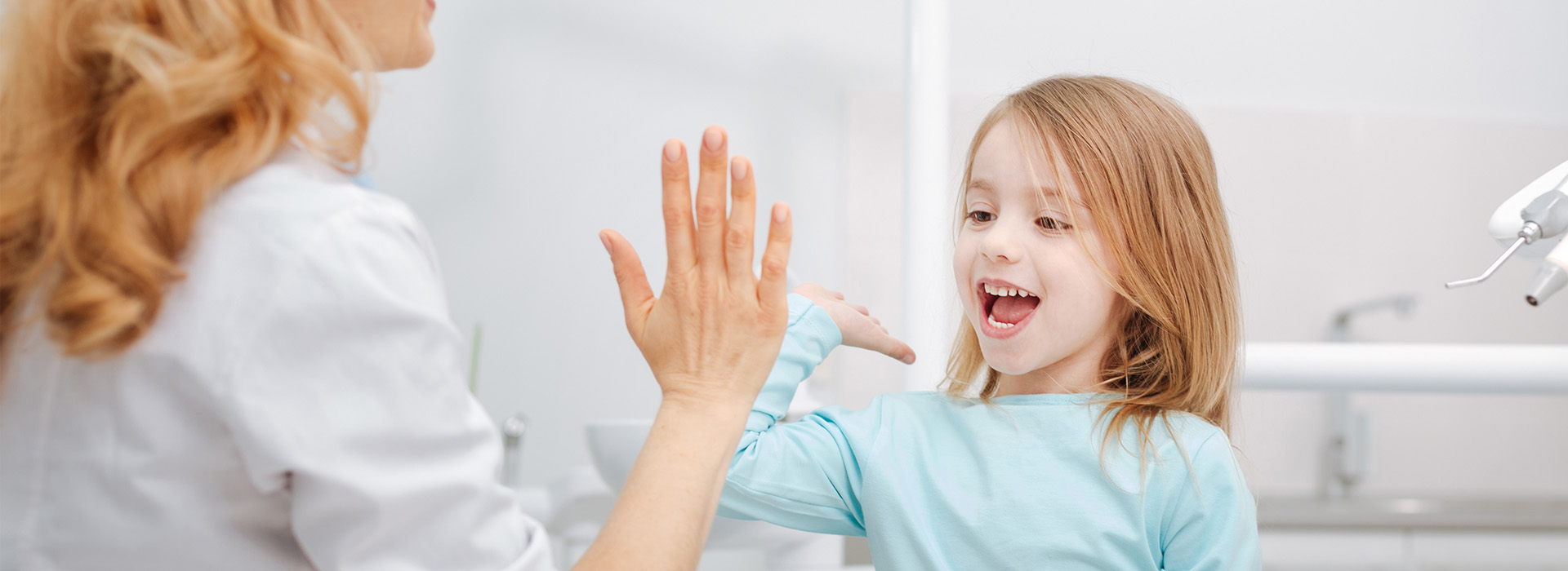 The image shows a young girl with blonde hair smiling broadly at the camera while standing next to an adult who appears to be giving her a high five, possibly in a bathroom setting.