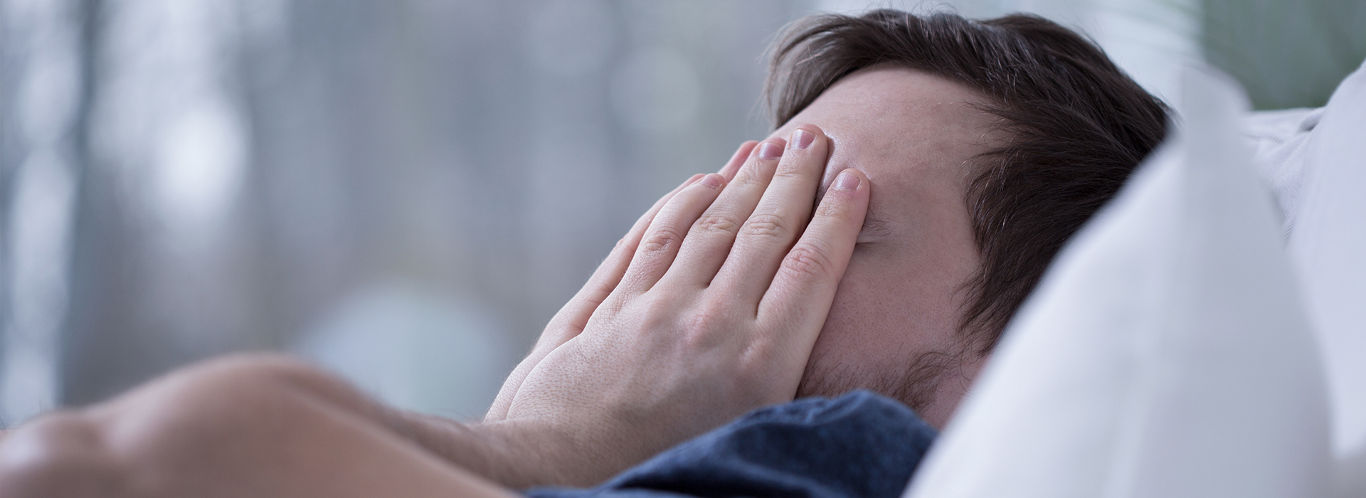 The image shows a man lying in bed with his face partially obscured by his hand, appearing to be in a state of distress or discomfort.