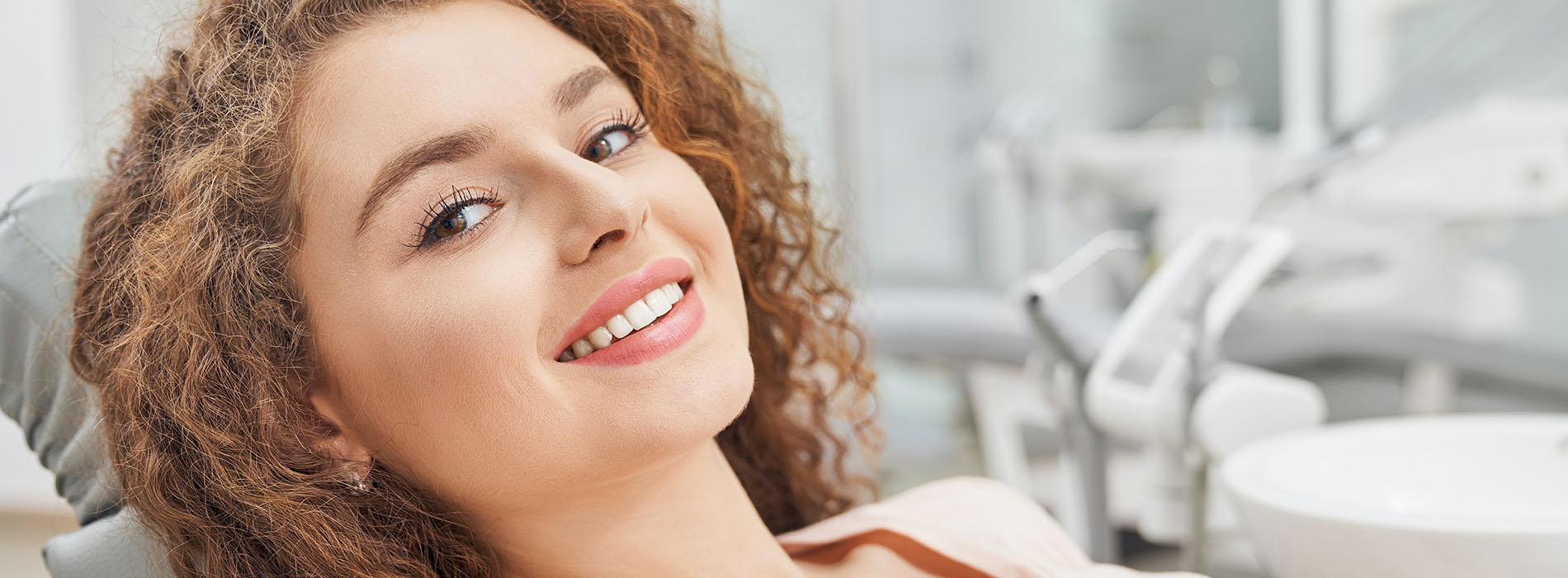 The image shows a smiling young woman with curly hair sitting in a dental chair, wearing a face mask, with her eyes closed, looking up towards the camera.